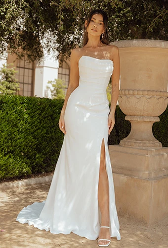 Bride in a minimalist white strapless wedding dress with a dramatic thigh slit, posing against a stone planter in an upscale garden venue