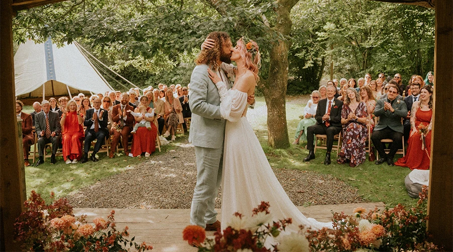 Real bride Georgie wearing an off-the-shoulder boho All Who Wander wedding dress while kissing her groom at the alter