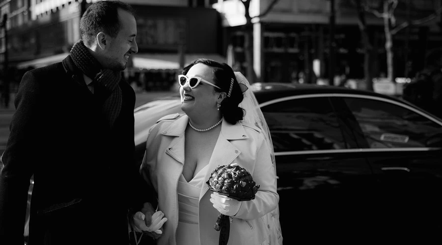 Real bride Priscilla wearing white cat-eye sunglasses and a leather jacket smiling with her groom Vincent.
