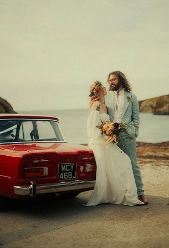 Real bride Georgie in a boho All Who Wander wedding dress posing with her groom and vintage red car near the ocean.