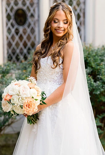 Real bride Janet Elizabeth holding a peach and white bouquet in a lace and sequin ballgown with a flowing veil.