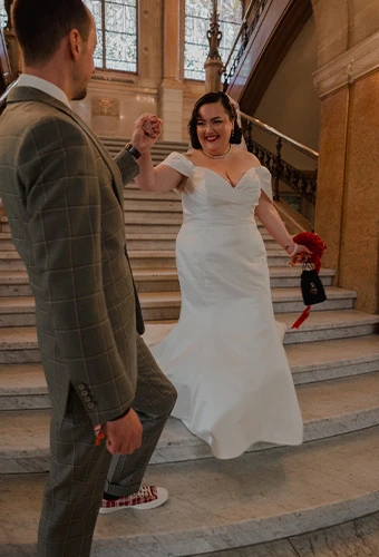 Real bride Priscilla holding hands with her groom on a grand staircase in an off-the-shoulder fit-and-flare gown.