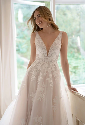 Woman wearing a blush-toned wedding dress with delicate floral lace details and a deep V-neckline, standing by a bright window