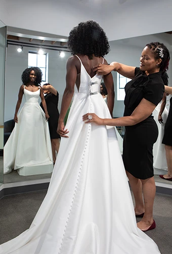 Bride tries on silk wedding dress while bridal stylist makes adjustments to the back.