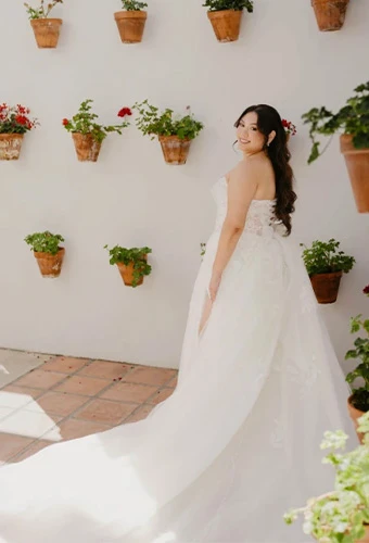 Real bride Maria in a white strapless wedding dress posing against a white wall decorated with potted flowers