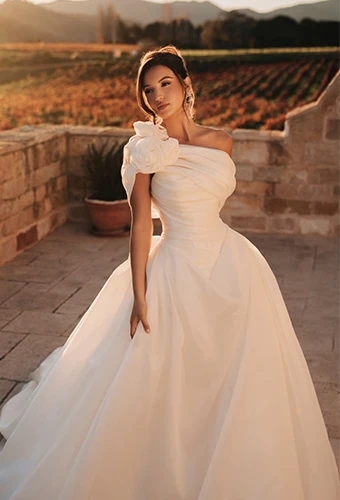 Bride wearing a-line wedding dress with asymmetrical one shoulder and floral accent posing on balcony