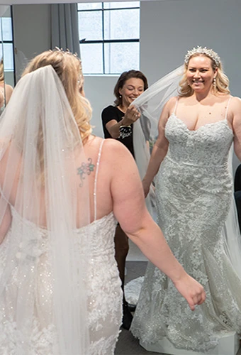 Bride-to-be trying on floral fit-and-flare wedding dress while bridal stylist adjusts veil