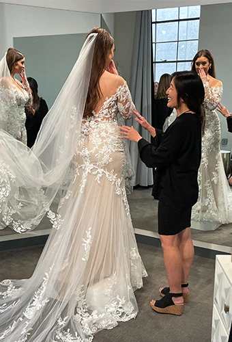 Bride-to-be tries on mermaid wedding gown while bridal stylists adjust sleeves and veil