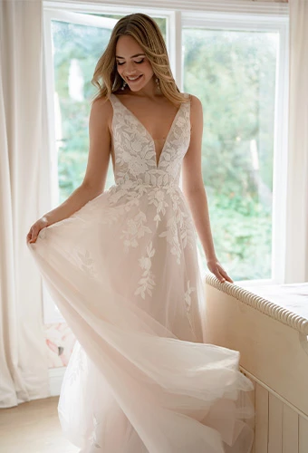 Woman wearing a blush-toned wedding dress with delicate floral lace details and a deep V-neckline, standing by a bright window