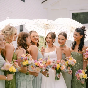 A bridal party celebrating outdoors, featuring a bride in a white simple satin gown and seven bridesmaids in mismatched shades of olive and sage green dresses, all holding colorful spring bouquets and paper parasols.