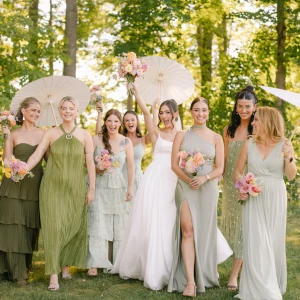 A smiling bridal party celebrating outdoors, with the bride raising her bouquet in a simple white slip dress with a side slit. Her seven bridesmaids wear a variety of styles in mixed olive and sage green shades, holding mixed spring bouquets and white paper parasols.