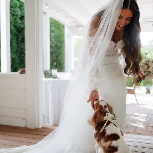 A smiling bride in a white long-sleeve lace and satin wedding dress reaches down to pet a Cavalier King Charles Spaniel dog wearing a white floral collar, on a rustic porch.