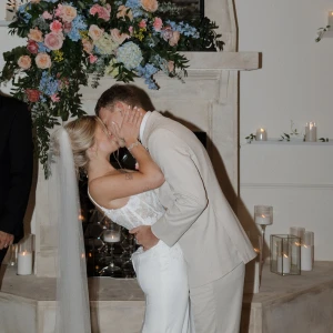A bride in a fitted, deep V-neck lace bodice wedding dress with a long veil kisses the groom, who is wearing a light tan suit, in front of a fireplace decorated with a large pink and blue floral arrangement.