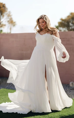A-Line wedding dress by Stella York, style SY8244. Showcases a strapless sweetheart neckline, detailed lace bodice, and a voluminous tulle skirt. Outdoor setting with green grass, pink wall, and trees.