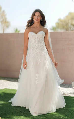 A-Line wedding dress by Stella York, style ID SY8205. Features a strapless sweetheart neckline, intricate lace bodice, and a flowing tulle skirt. Modeled outdoors on green grass with a soft pink wall backdrop.