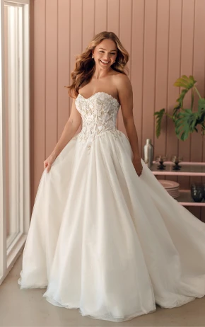 Ballgown wedding dress by Stella York, style SY8181. Includes a strapless sweetheart neckline, lace bodice, and a full tulle skirt. Outdoor setting with green grass, pink wall, and trees.