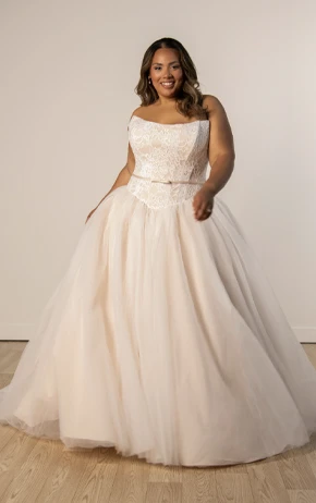 Stella York SY8189+ ballgown with a strapless sweetheart neckline, lace bodice, and full tulle skirt adorned with lace appliqués. Model poses outdoors on grass with a pink wall and trees.