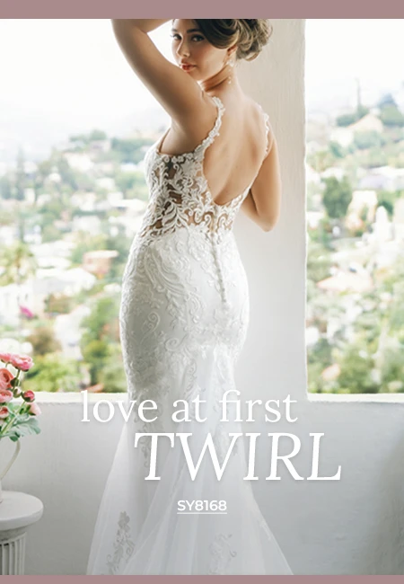 Stella York bridal gown SY8168, a mermaid wedding dress featuring an illusion lace bodice, deep open back, and intricate all-over lace and tulle skirt. The bride stands in a window overlooking a sunny cityscape.